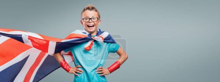 Téléchargez les photos : Mignon garçon souriant superhéros posant avec des bras akimbo et portant un drapeau britannique comme une cape, l'apprentissage des langues concept - en image libre de droit