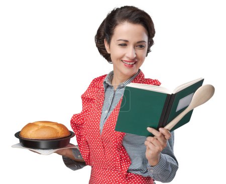 Photo for Smiling retro housewife holding a cookbook and a baking tin with a homemade cake - Royalty Free Image