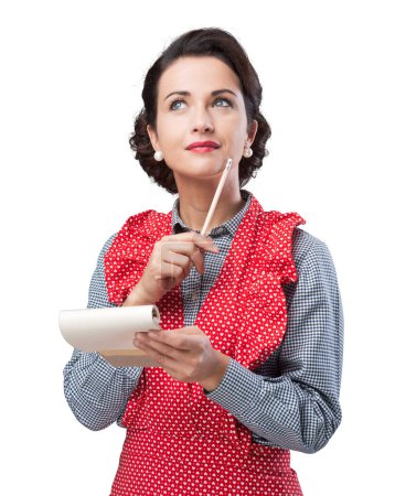 Photo for Attractive smiling housewife thinking and writing down a shopping list on a notepad - Royalty Free Image