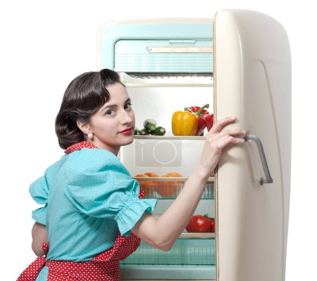 Femme au foyer de style vintage dans la cuisine préparant le déjeuner, elle ouvre le réfrigérateur et regarde la caméra