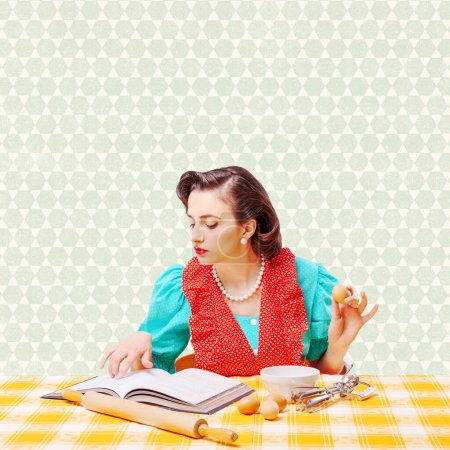 Téléchargez les photos : Belle ménagère de style vintage préparant un dessert à la maison, elle vérifie la recette dans un livre de cuisine et mélange des ingrédients dans un bol - en image libre de droit