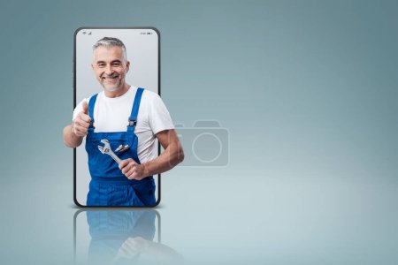 Foto de Cheerful repairman and plumber giving a thumbs up in a smartphone videocall and smiling, online  service concept - Imagen libre de derechos