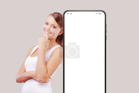 Téléchargez les photos : Jeune femme souriante appuyée contre un grand smartphone avec écran vierge - en image libre de droit