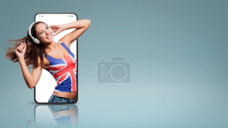 Téléchargez les photos : Adolescente souriante portant un débardeur drapeau anglais, elle écoute de la musique avec des écouteurs et danse dans un vidéocall smartphone - en image libre de droit