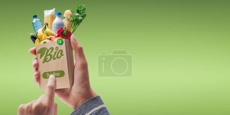 Foto de Customer holding a miniature shopping bag and ordering organic grocery online, she is pressing the buy now button - Imagen libre de derechos
