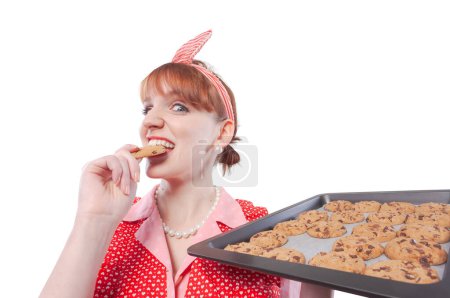 Téléchargez les photos : Joyeuse femme au foyer de style vintage dégustant ses bonbons maison fraîchement cuits, elle tient une plaque de cuisson pleine de biscuits - en image libre de droit