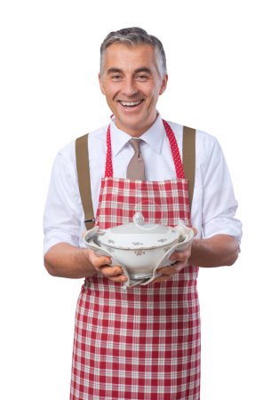 Photo for Handsome smiling man in apron serving dinner in a tureen - Royalty Free Image