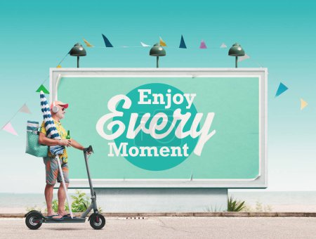Photo for Happy senior man riding an electric scooter and going to the beach, motivational poster in the background: enjoy every moment - Royalty Free Image