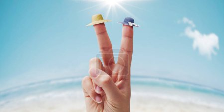 Photo for Hand making a V sign and wearing hats on two fingers, beach in the background, summer vacations concept - Royalty Free Image