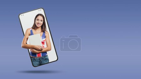Téléchargez les photos : Jeune fille souriante tenant un livre relié dans un vidéocall smartphone et souriant, concept d'apprentissage des langues et d'éducation - en image libre de droit