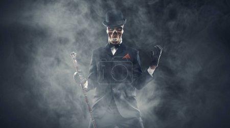 Téléchargez les photos : Personnage maléfique effrayant avec tête de crâne portant un costume élégant, il regarde la caméra et pose entouré de brouillard, concept d'horreur - en image libre de droit