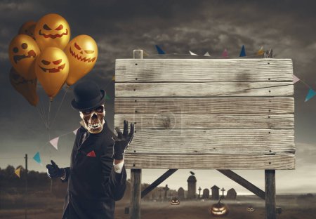 Photo for Creepy Halloween skeleton character holding balloons and showing a wooden sign: festivity and celebration concept - Royalty Free Image