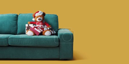 Niedlicher Teddybär mit Weihnachtsmütze auf der Couch und vielen Geschenken, Feiertagen und Feierlichkeiten