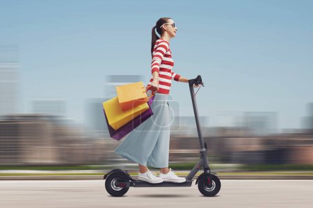 Photo for Happy young woman carrying many shopping bags and riding an electric scooter, cityscape in the background, sale and shopping concept - Royalty Free Image