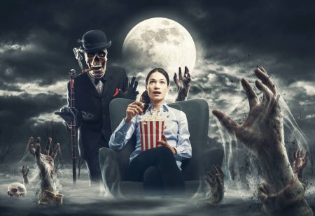 Foto de Mujer asustada viendo películas de terror y comiendo palomitas de maíz, ella está rodeada de espeluznantes manos de zombies y un monstruo esqueleto está de pie detrás de ella - Imagen libre de derechos