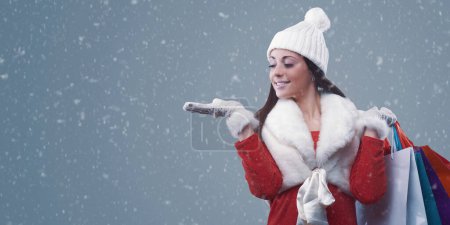 Foto de Mujer feliz haciendo compras de Navidad bajo la nieve, ella está sonriendo y sosteniendo muchas bolsas - Imagen libre de derechos
