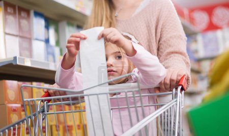 Disappointed girl at the supermarket with her mother, she is holding a long expensive grocery receipt