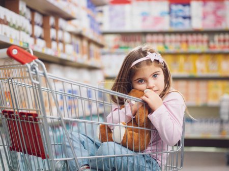 Téléchargez les photos : Portrait d'une jolie petite fille au supermarché, elle est assise à l'intérieur d'un chariot et serre son ours en peluche - en image libre de droit