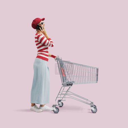 Foto de Mujer feliz teniendo una llamada telefónica y empujando un carro de supermercado vacío, longitud completa - Imagen libre de derechos