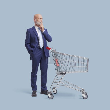 Photo for Pensive doubtful businessman standing behind a shopping cart, he is thinking with hand on chin - Royalty Free Image