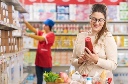 Foto de Mujer haciendo compras de comestibles en el supermercado y comprobando una etiqueta de alimentos en un producto, empleado de stock trabajando en el fondo - Imagen libre de derechos