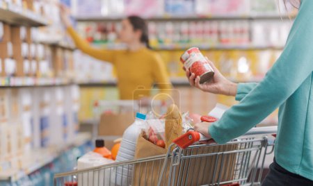 Foto de Mujer poniendo artículos en el carrito de la compra en la tienda de comestibles, ella está sosteniendo una lata y leyendo la etiqueta - Imagen libre de derechos