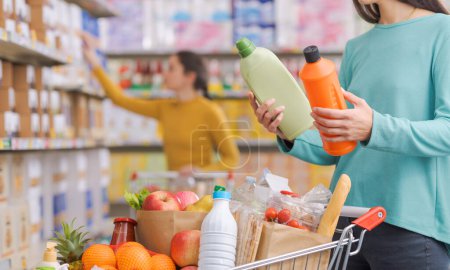Foto de Mujer comprando productos en la tienda de comestibles, está comparando dos botellas de detergentes - Imagen libre de derechos