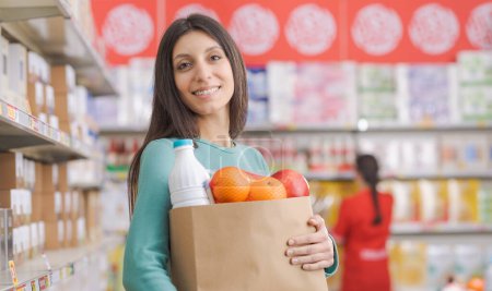 Photo for Smiling young woman holding a bag full of groceries and looking a camera, stock clerk working and supermarket shelves in the background - Royalty Free Image