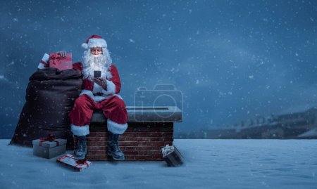 Téléchargez les photos : Le Père Noël livre des cadeaux la veille de Noël, il est assis sur une cheminée et se connecte en ligne avec son smartphone - en image libre de droit
