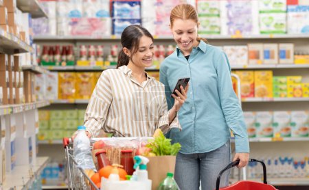 Foto de Sonriendo a las mujeres en el supermercado comprando comestibles juntos, una mujer está mostrando su teléfono inteligente a su amigo, concepto de compras de comestibles - Imagen libre de derechos