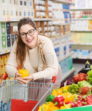 Foto de Mujer sonriente empujando un carrito de la compra y la compra de verduras frescas y saludables en el supermercado, compras de comestibles y concepto de nutrición - Imagen libre de derechos