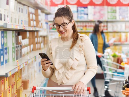 Téléchargez les photos : Une cliente heureuse faisant des courses au supermarché et utilisant son smartphone - en image libre de droit