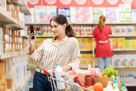 Téléchargez les photos : Femme faisant des courses au supermarché, elle vérifie les informations sur le produit à l'aide d'une application scanner sur son smartphone - en image libre de droit