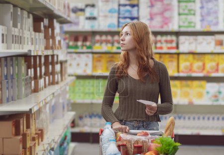 Téléchargez les photos : Jeune femme faisant des courses au supermarché, elle pousse un chariot plein et tient une liste de courses - en image libre de droit