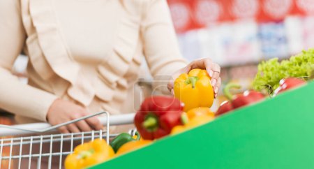 Téléchargez les photos : Femme poussant un chariot et achetant des légumes frais au supermarché, elle cueille un poivron - en image libre de droit