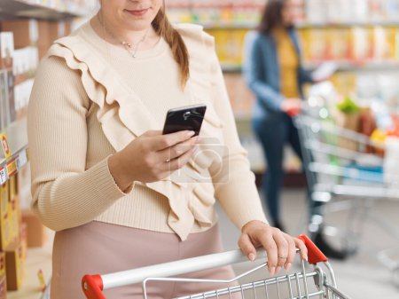 Photo for Customer doing grocery shopping at the supermarket and using her smartphone - Royalty Free Image