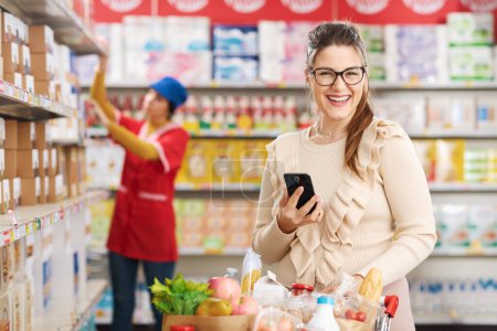 Photo for Smiling woman buying groceries and using a smartphone at the grocery store - Royalty Free Image