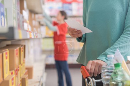 Foto de Cliente compra comestibles en el supermercado, ella está sosteniendo una lista de supermercados y empujando un carrito de compras completo - Imagen libre de derechos