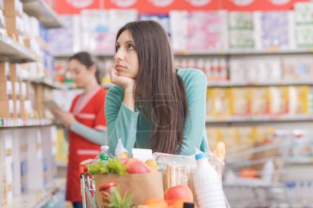 Foto de Mujer joven aburrida apoyada en el carrito de la compra en la tienda de comestibles, ella está mirando a su alrededor y sintiéndose cansada - Imagen libre de derechos