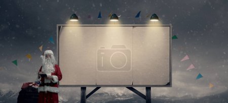 Photo for Santa Claus checking a list and delivering gifts, he is standing next to a billboard sign with blank posters - Royalty Free Image