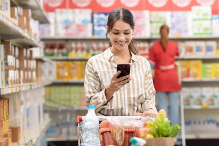 Foto de Mujer sonriente empujando un carrito de compras completo y utilizando un teléfono inteligente en el supermercado, compras de comestibles y el concepto de tecnología - Imagen libre de derechos