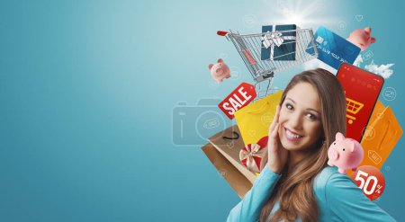 Foto de Joven alegre soñando con las compras en línea, ella está rodeada de artículos de compras y sonriendo - Imagen libre de derechos
