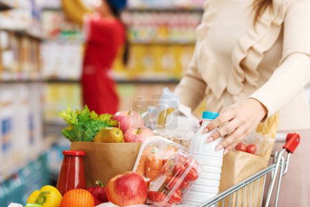 Téléchargez les photos : Femme achetant des produits frais au supermarché, elle pousse un panier plein et tient une bouteille de lait - en image libre de droit