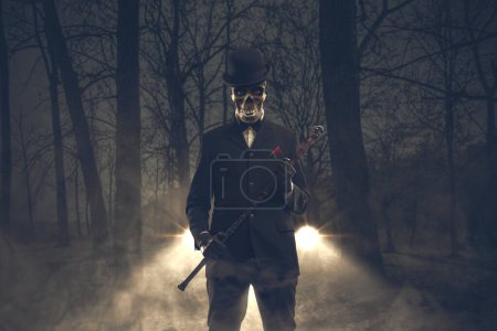 Téléchargez les photos : Démon maléfique effrayant avec tête de crâne portant un costume élégant et tenant un bâton de marche, il se tient dans la forêt sombre, l'horreur et le concept d'Halloween - en image libre de droit