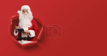 Téléchargez les photos : Père Noël pensif écrivant une liste sur un bloc-notes et surgissant d'un trou dans le papier, espace de copie vierge - en image libre de droit