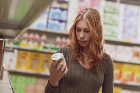 Foto de Mujer joven haciendo compras de comestibles en el supermercado, que está sosteniendo una lata y comprobar la información en la etiqueta del producto - Imagen libre de derechos