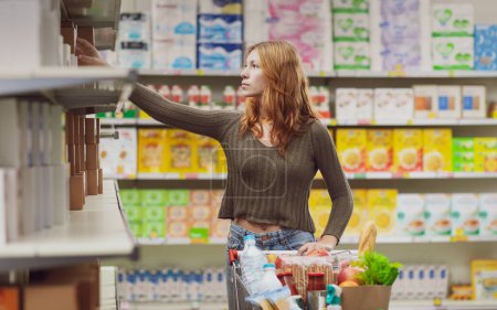 Téléchargez les photos : Femme faisant des courses au supermarché, elle prend un produit de l'étagère du magasin - en image libre de droit
