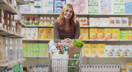 Téléchargez les photos : Portrait d'une jeune femme au supermarché, elle s'appuie sur la poignée du chariot et sourit à la caméra - en image libre de droit