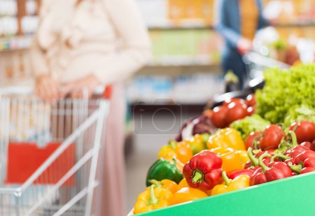 Foto de Verduras frescas en la sección de productos en el supermercado y mujer empujando un carrito de compras - Imagen libre de derechos