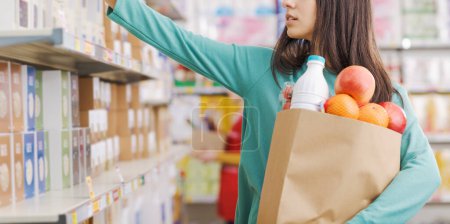 Foto de Mujer atractiva joven tomando productos de la estantería en la tienda de comestibles, ella está sosteniendo una bolsa de compras completa - Imagen libre de derechos